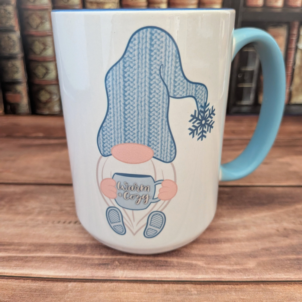 A white mug with a blue handle. The image on the mug is a winter gnome in a blue knit hat, holding a blue coffee mug with the words"Warm & Cozy."