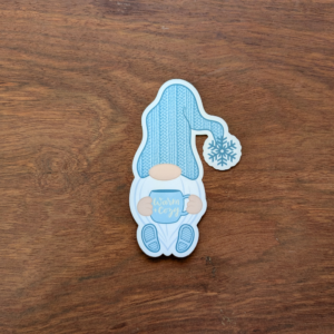 A winter gnome sticker wearing a blue knit hat and holding a blue coffee mug. The mug reads "Warm and Cozy."