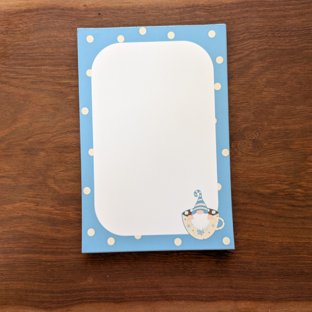 A blue and white polka-dot notepad with a winter gnome in the bottom right corner. The gnome is wearing a blue striped hat and sitting in a cup.