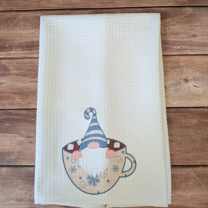A white waffle towel with a winter gnome image at the bottom. The gnome is wearing a blue-and-cream striped hat and is sitting in a cream cup filled with hot chocolate.