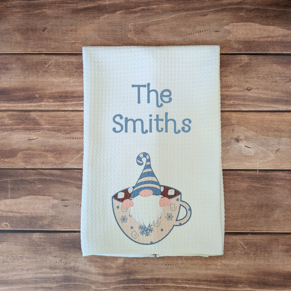 A personalized white waffle kitchen towel with the words "The Smiths." A gnome sitting in a cream cup decorated with blue snowflakes is under the text.