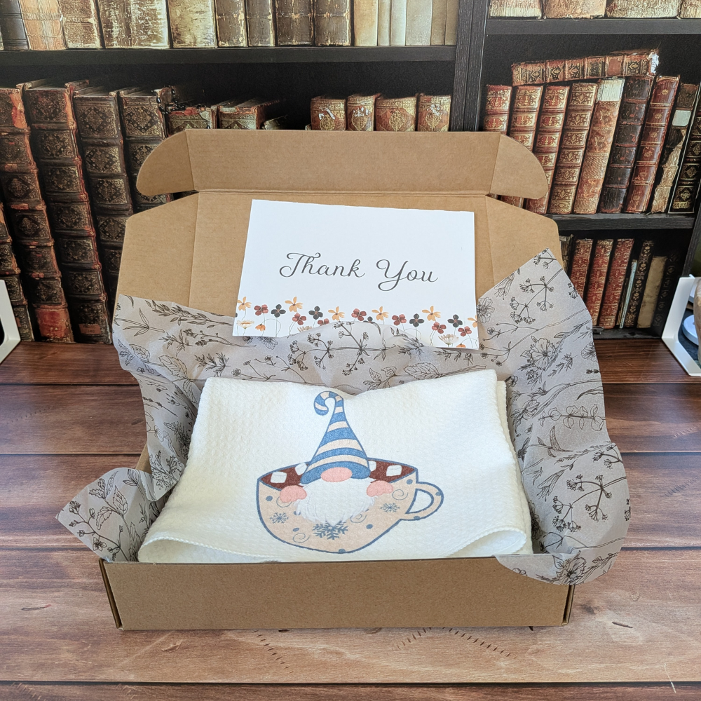 A brown box with a white waffle towel inside. The winter gnome is on the towel, and a thank-you note is above the towel.